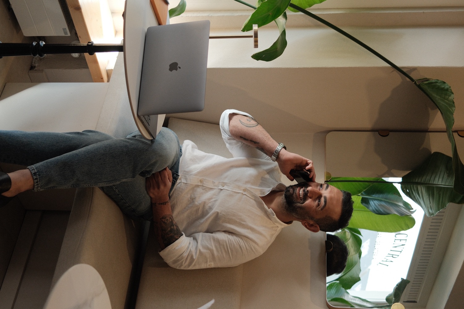 Emrah Olgac sitting in a bright, plant-filled café, smiling during a phone call with a laptop beside him
